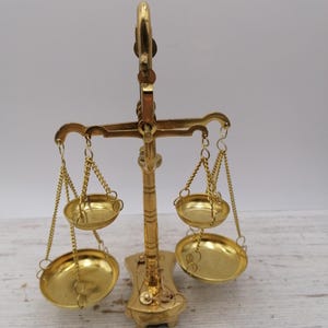 Peut inclure: Une balance dorée avec deux plateaux suspendus et un support central. La balance est en métal et présente un design décoratif. Les plateaux sont ronds et suspendus par des chaînes.