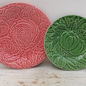 Peut inclure: Deux assiettes rondes en céramique avec des motifs en relief. La plus grande assiette est rose et présente un motif dense de roses épanouies et de feuilles. La plus petite assiette est verte et affiche un motif de fruit entouré de feuillage.
