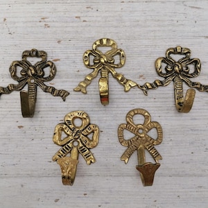 May include: Five gold-toned metal hooks shaped like bows. Each hook has a decorative design with a bow shape and a loop at the top for hanging items.