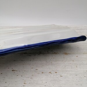 May include: A rectangular white ceramic serving dish with a blue trim. The dish has a slightly raised, ridged surface and a curved edge. The background is a light-coloured, textured surface.