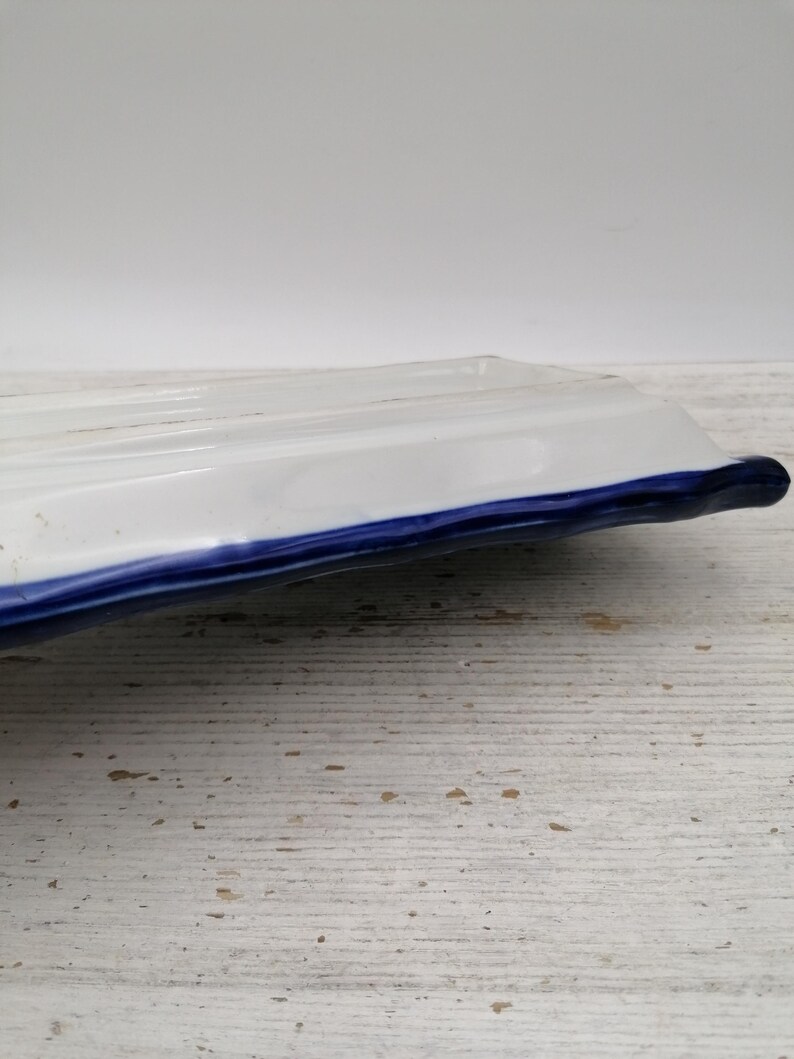May include: A rectangular white ceramic serving dish with a blue trim. The dish has a slightly raised, ridged surface and a curved edge. The background is a light-coloured, textured surface.