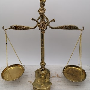 Könnte beinhalten: Eine antike Messingwaage mit zwei ovalen Pfannen, die an Ketten aufgehängt sind. Die Waage weist aufwendige Details auf, darunter delfinförmige Arme und eine dekorative Oberseite. Die Waage ist goldfarben und steht auf einer Holzoberfläche.
