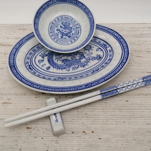May include: A white and blue porcelain Asian-style dining set. The set includes a bowl, an oval plate, a pair of chopsticks, and a chopstick rest. The plate and bowl feature a blue dragon design. The chopsticks have a blue geometric pattern.