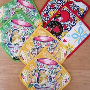 Könnte beinhalten: Eine Sammlung quadratischer Topflappen aus Stoff mit aufgedruckten Motiven von gestapelten Teetassen, kleinen Vögeln und Löffeln. Das Set enthält grün, gelb und rot eingefasste Stücke mit floralen und geometrischen Mustern in bunten Farben.