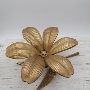 May include: A gold-colored metal flower sculpture with six petals and two leaves. The center of the flower has two small, colorful gems. The sculpture is likely a decorative item.