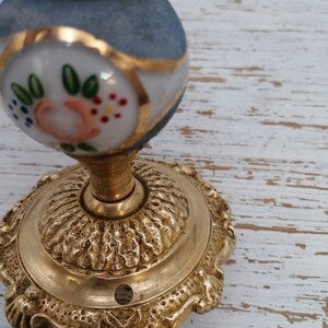 May include: A gold-toned door knob with a blue and white porcelain top. The porcelain top has a floral design with pink, green, and white flowers. The knob has a decorative base with a scalloped edge.