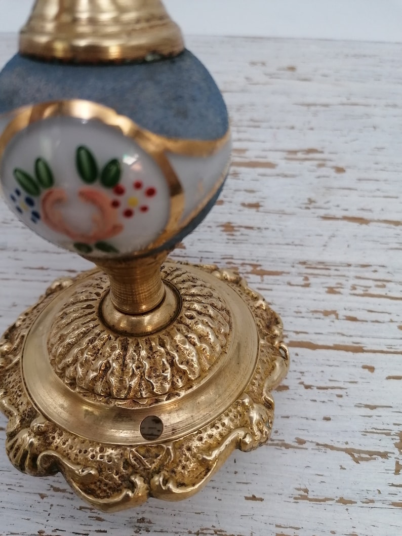 May include: A gold-toned door knob with a blue and white porcelain top. The porcelain top has a floral design with pink, green, and white flowers. The knob has a decorative base with a scalloped edge.