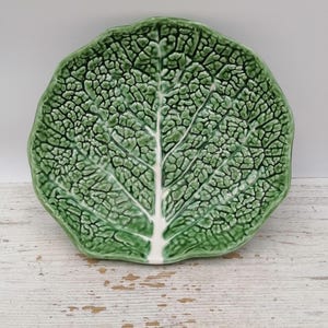 Może przedstawiać: Ceramiczny talerz w kształcie zielonego liścia kapusty z błyszczącym wykończeniem. Powierzchnia posiada bardzo szczegółową, tłoczoną fakturę imitującą żyłki i pomarszczony wygląd prawdziwego liścia, z wyraźną białą łodygą pośrodku.