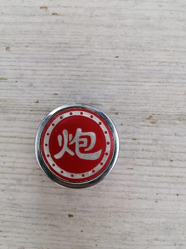 Puede incluir: Un bot&oacute;n redondo plateado y rojo con el car&aacute;cter chino para "fuego" en blanco sobre fondo rojo. El bot&oacute;n tiene un borde blanco punteado.
