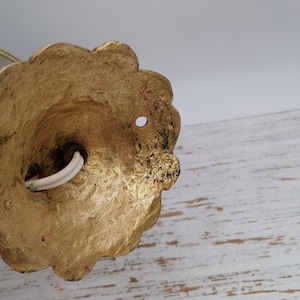 May include: Close-up of the back of a gold-colored wall sconce with a decorative, scalloped edge. The sconce has a hole in the center and a white electrical cord coming out of it.
