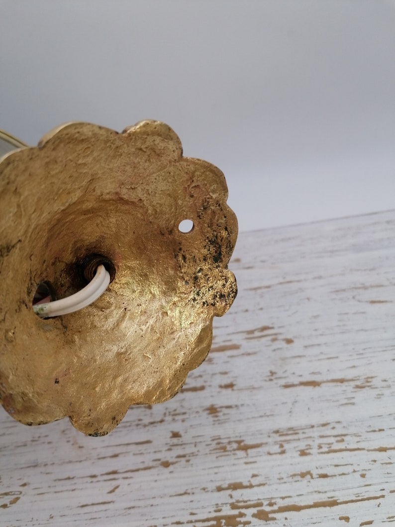 May include: Close-up of the back of a gold-colored wall sconce with a decorative, scalloped edge. The sconce has a hole in the center and a white electrical cord coming out of it.