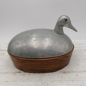 May include: A decorative duck-shaped box. The lid is made of silver-colored metal, and the eyes are red. The base of the box is made of wood. The duck's head is upright, and the beak is pointed.
