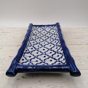 May include: A rectangular ceramic serving dish with a bamboo-style frame in glossy dark blue. The interior features a white background with a repeating blue floral pattern. The dish is designed for serving sushi or appetizers.