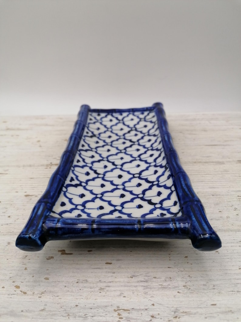 May include: A rectangular ceramic serving dish with a bamboo-style frame in glossy dark blue. The interior features a white background with a repeating blue floral pattern. The dish is designed for serving sushi or appetizers.