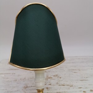 May include: A small table lamp with a dark green fabric shade and a gold trim. The lamp has a white ceramic base and a gold metal stem.