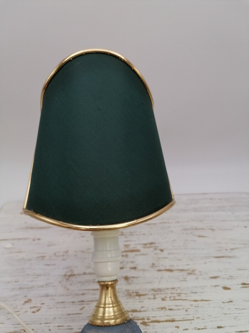 May include: A small table lamp with a dark green fabric shade and a gold trim. The lamp has a white ceramic base and a gold metal stem.