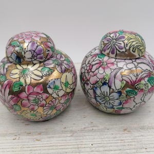 Op de afbeelding: Twee kleine, ronde porseleinen potten met bloemenmotieven. De potten zijn versierd met roze, paarse, gele, groene en gouden bloemen op een witte achtergrond.