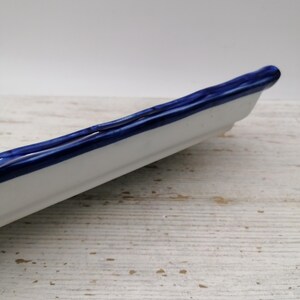 May include: A white ceramic serving dish with a deep blue rim. The dish is long and narrow, suitable for serving appetizers or small portions. The blue rim provides a decorative element to the simple design. The dish is approximately 25 cm long.