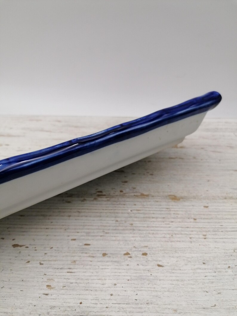 May include: A white ceramic serving dish with a deep blue rim. The dish is long and narrow, suitable for serving appetizers or small portions. The blue rim provides a decorative element to the simple design. The dish is approximately 25 cm long.