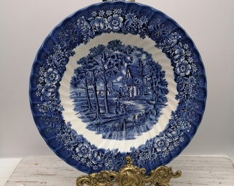 Op de afbeelding: Een blauw en wit keramiek bord met een bloemige rand en een scène met een huis en bomen in het midden. Het bord staat op een gouden metalen standaard.