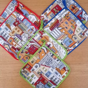 Puede incluir: Tres agarradores de tela cuadrados con una colorida ilustración de una calle europea con edificios en rojo, amarillo y azul. Cada agarrador tiene un borde distintivo en rojo, azul o verde e incluye una trabilla de tela a juego para colgar.