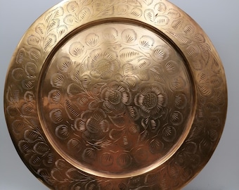 May include: A round, antique brass plate with intricate floral and leaf engravings. The plate is displayed on a decorative brass stand. The design features detailed patterns of flowers and foliage, creating a vintage aesthetic.