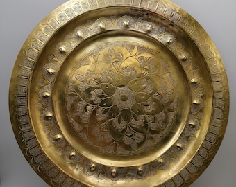 May include: A large, round, brass serving tray with intricate floral designs etched into the surface. The tray has a raised rim with a scalloped edge and is supported by a decorative metal stand.