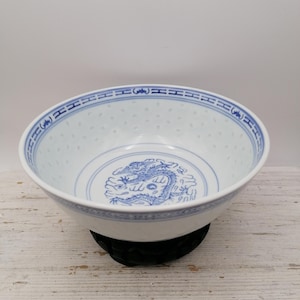 May include: White porcelain bowl with a blue dragon design in the center. The bowl has a blue rim with a repeating pattern and small holes around the sides. It sits on a black wooden stand.