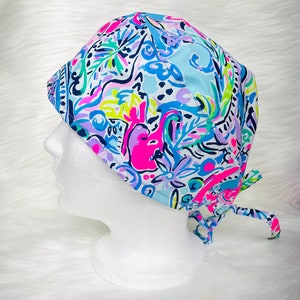 Könnte beinhalten: Eine bunte OP-Haube mit einem lebendigen Blumen- und abstrakten Muster in Pink-, Blau-, Grün- und Lilatönen. Die Haube hat einen Bindverschluss am Rücken und wird auf einem weißen Schaufensterkopf präsentiert. Der Stoff hat einen hellblauen Hintergrund.