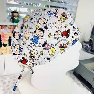 May include: A light gray surgical scrub cap featuring a repeating pattern of cartoon characters, including Snoopy, Charlie Brown, and Lucy. The cap has ties for a secure fit and is displayed on a white mannequin head.