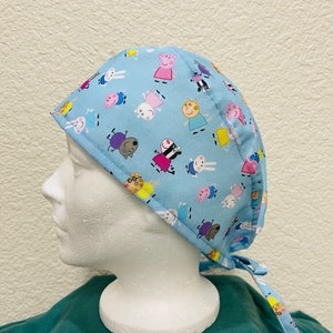 May include: A light blue surgical scrub cap featuring a repeating pattern of cartoon characters. The cap is tied at the back with matching fabric ties. The characters are in various colors, including pink, yellow, and purple.