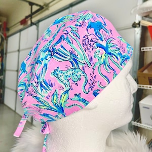 May include: A pink surgical scrub cap with a colorful pattern of animals and foliage in blue, green, and white. The cap has ties for a secure fit. The cap is displayed on a white mannequin head.