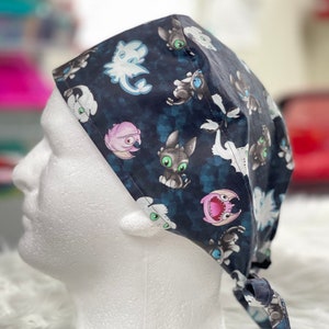 May include: A dark blue surgical scrub cap featuring a repeating pattern of cartoon dragons in various colors. The cap is tied at the back and is displayed on a white mannequin head.