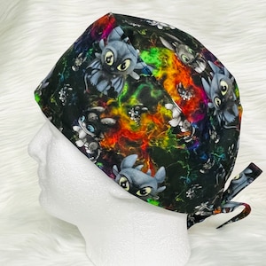 May include: A black surgical scrub cap featuring a colorful design with images of a cartoon dragon. The cap has a tie closure and is displayed on a white mannequin head. The background is a soft, white surface.