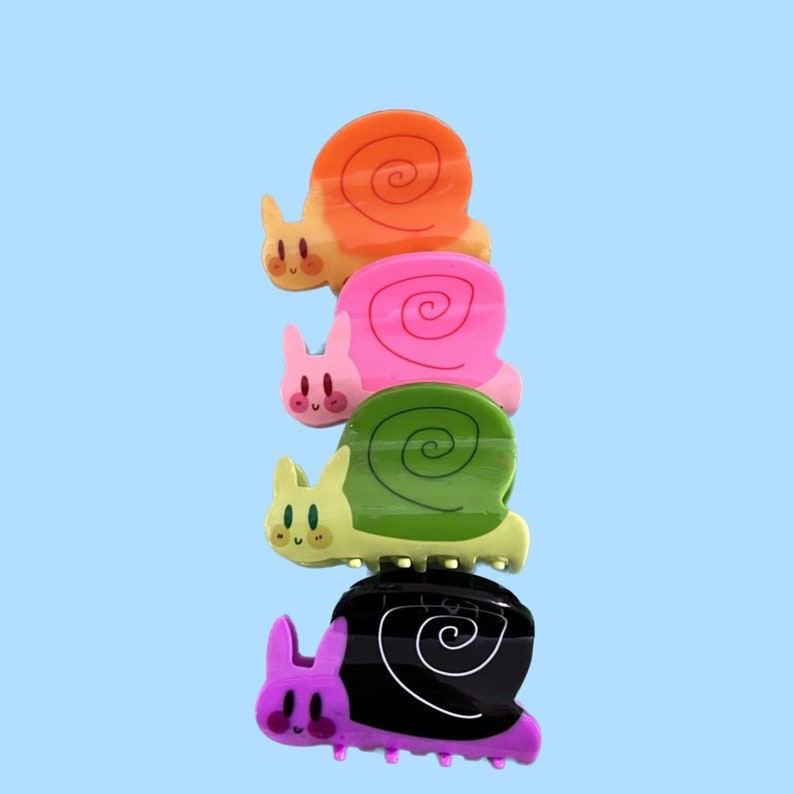 May include: Four colorful snail-shaped hair clips. The clips are stacked on top of each other, starting with orange, then pink, green, and finally black. Each clip has a cute cartoon face and a spiral shell.