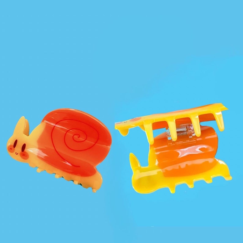 May include: Two plastic hair clips shaped like snails. One is orange and yellow with a red swirl on the shell. The other is yellow with a clear plastic clasp.