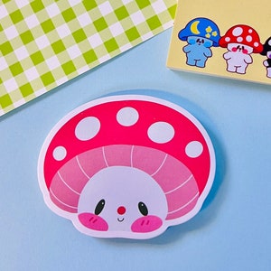 Może przedstawiać: Naklejka w kształcie czerwonego i białego grzyba z różową i białą spodem. Grzyb ma białą twarz z czarnymi oczami i czerwonym nosem. Uśmiecha się.