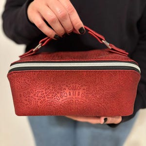 May include: A red cosmetic bag with a textured, patterned surface and a silver zipper. The bag has a matching red handle and is held by a person. The bag is approximately 8 inches long, 6 inches wide, and 4 inches tall.