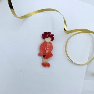 Puede incluir: Un broche de plástico rojo y blanco con forma de niña con el pelo rojo rizado. La niña lleva un vestido rojo y tiene la cara blanca.