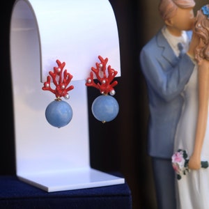 May include: A pair of earrings with red coral branches and blue round stones. The earrings are displayed on a white stand.