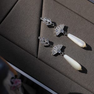 May include: A pair of silver earrings with a fan-shaped design and a white pearl drop. The earrings are on a gray surface.