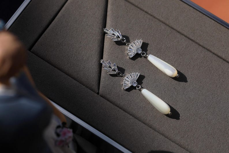 May include: A pair of silver earrings with a fan-shaped design and a white pearl drop. The earrings are on a gray surface.