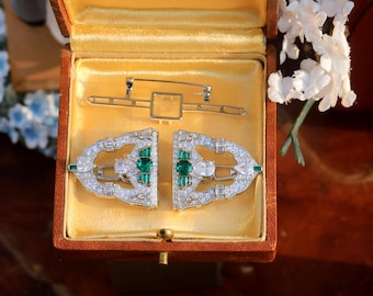 May include: A pair of silver brooches with emerald green and clear stones, displayed in a velvet-lined box.