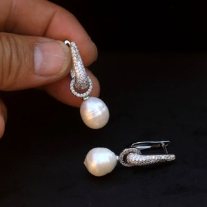 Pode incluir: Brincos pendurados com grandes pérolas brancas em forma de gota. Os brincos têm um design em metal prateado com um topo em forma de cone e um anel de pequenas pedras transparentes. Em um fundo escuro.