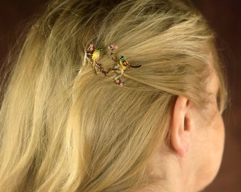 May include: A gold hair clip with two yellow birds perched on a branch with pink flowers. The birds have green eyes and black accents.