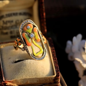 May include: A gold ring featuring a flower design. The flower is crafted from enamel and displays a yellow, green, and orange colour scheme. The ring is set with small diamonds.