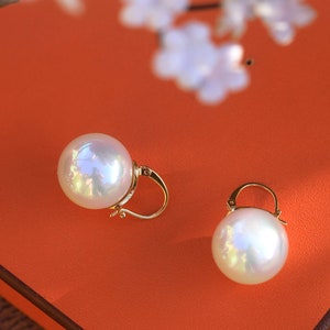 May include: A pair of gold pearl earrings with a large, round, white pearl on each earring. The earrings are on a bright orange surface.