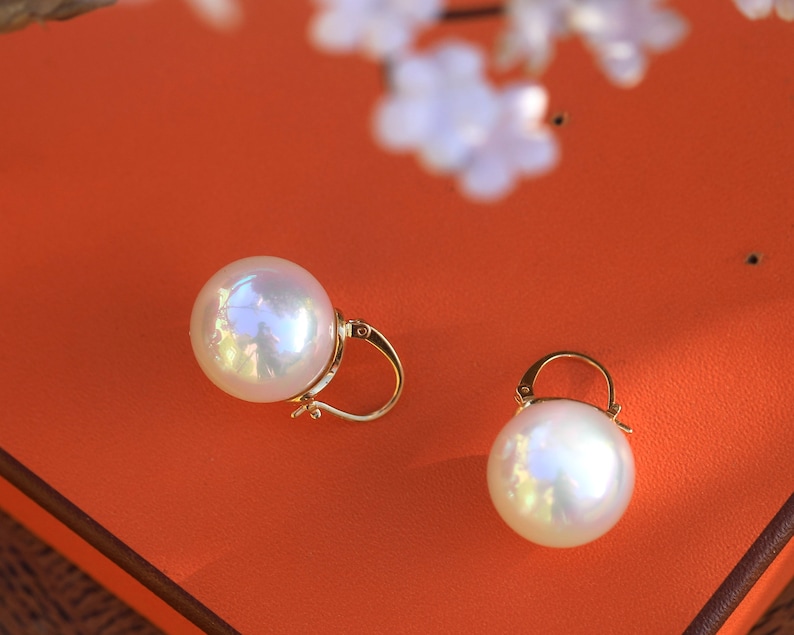May include: A pair of gold pearl earrings with a large, round, white pearl on each earring. The earrings are on a bright orange surface.