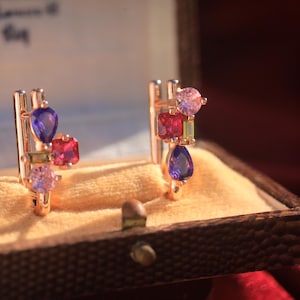 May include: A pair of gold-toned earrings with colorful gemstones, including purple, red, and pink stones. The earrings are displayed in a vintage jewelry box with a soft, yellow interior. The gemstones are cut in various shapes.