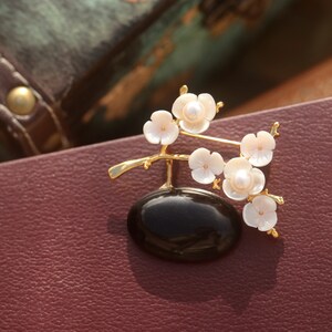 May include: A gold-toned brooch featuring a black oval base with a delicate branch of white flower blossoms. Each blossom has a pearl center. The brooch is set against a burgundy leather surface.